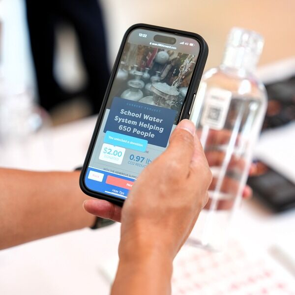 A person scanning a QR code on a water bottle with a smartphone screen showing  'School water system helping 650 people'.